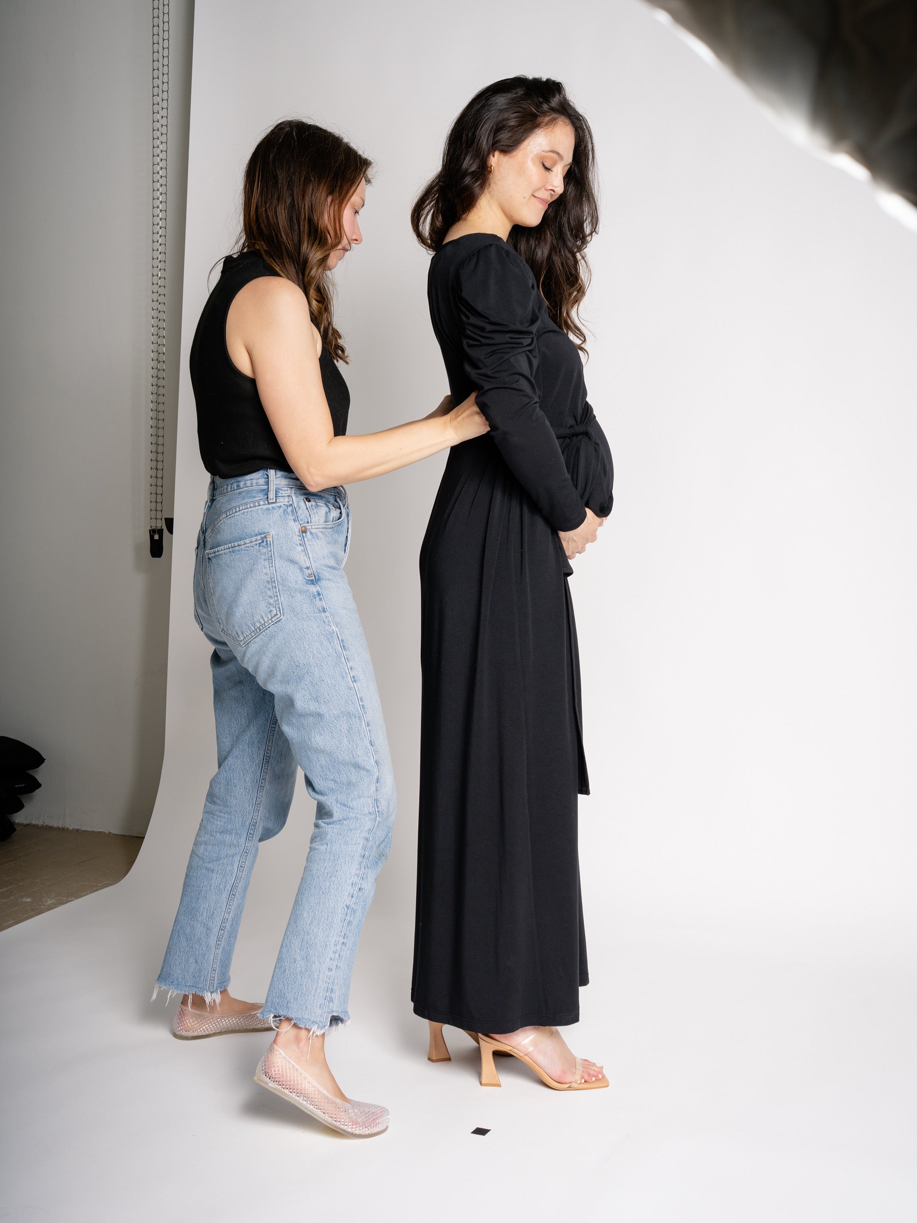 Two women standing on a white floor, one wearing a black dress and the other in a black top and jeans.