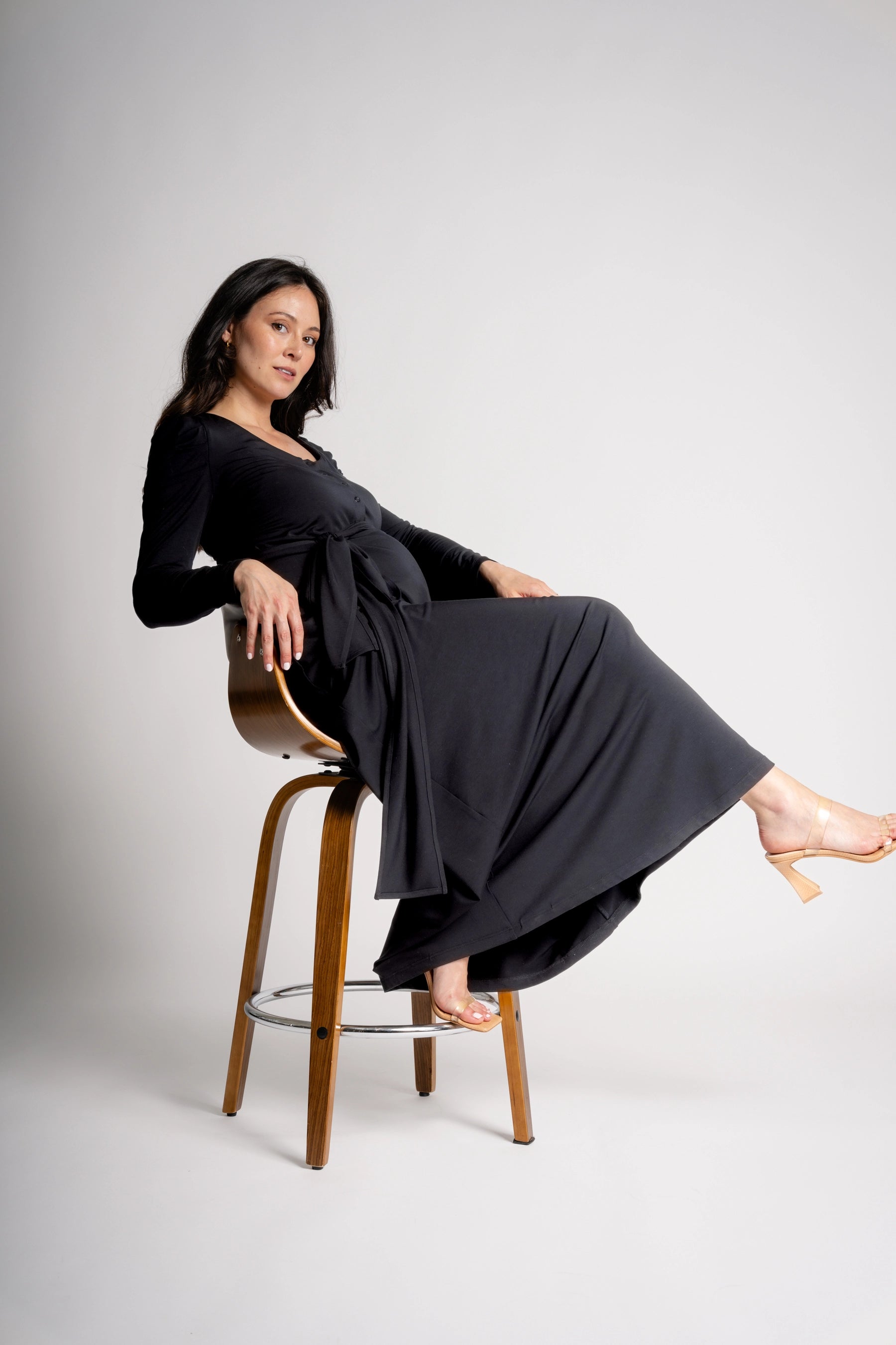 Woman in a black dress sitting on a wooden stool against a gray background
